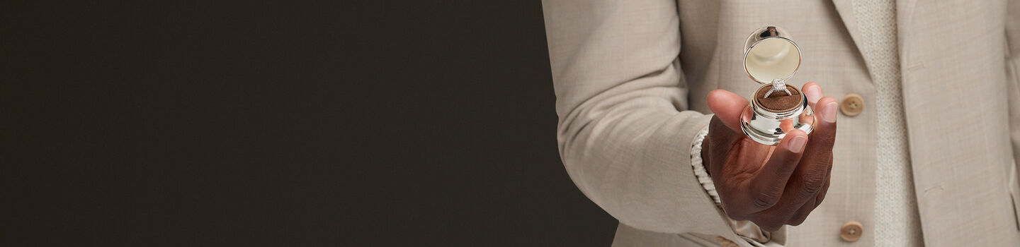 Birks engagement ring displayed in a silver bell box, held by a man in a beige suit.